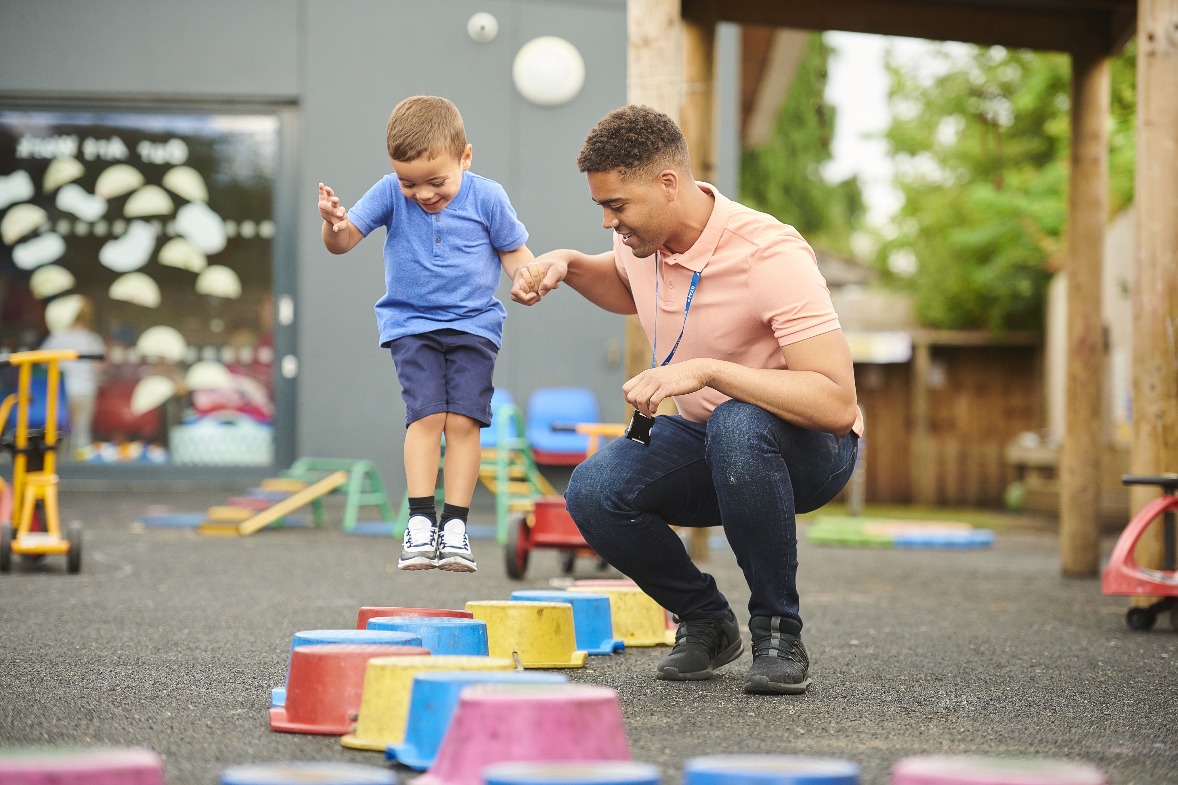 Boy and male teacher playing outdoors, jumping and balancing on colourful plastic buckets arranged in a line